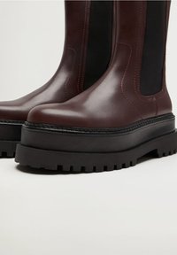 Bottines en cuir bourgogne avec des panneaux élastiques noirs, semelle épaisse noire et motif de crampons prononcé. Texture lisse sur l'ensemble.