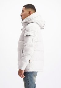 Lichtgrijze pufferjack met een afneembare capuchon, ritssluiting aan de voorkant en een geborduurde textuur. Beschikt over zijzakken en een kleuraccent op de zoom.