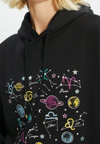 Sweat à capuche noir arborant des imprimés colorés de signes du zodiaque et d'images célestes, incluant des planètes, des étoiles et des constellations dans diverses teintes.