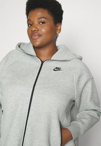 Nike pelēks zip-up džemperis ar kapuci, ar melniem akcentu šuvēm un logotipu, izgatavots no mīkstas, teksturētas auduma. Iekļautas sānu kabatas.