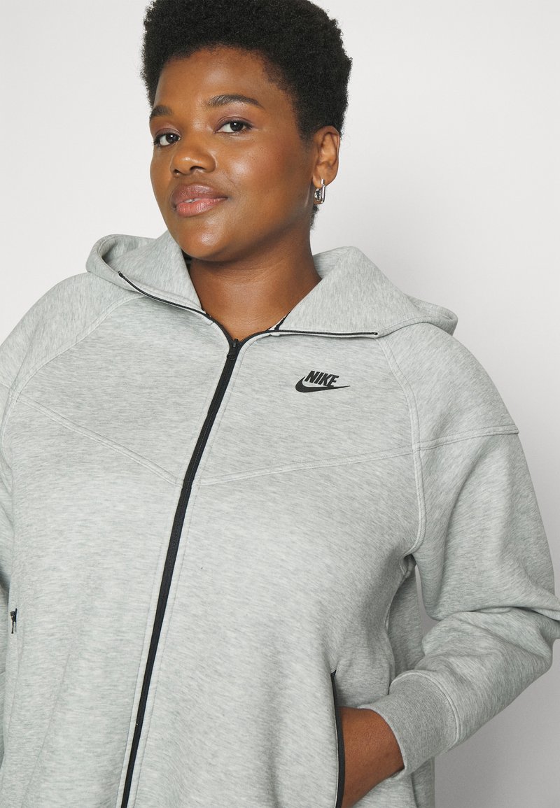 Haut à capuche zippé gris Nike, avec des coutures et un logo noirs, fabriqué en tissu doux et texturé. Poches latérales incluses.