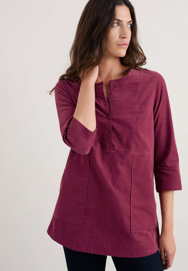 Túnica burdeos de botones con mangas de tres cuartos, dobladillo curvado, dos bolsillos frontales y tejido texturizado. Diseño simple y casual.