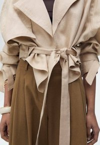 Veste beige ceinturée avec des manches surdimensionnées, associée à un pantalon large marron taille haute et à des bracelets en or au poignet gauche.