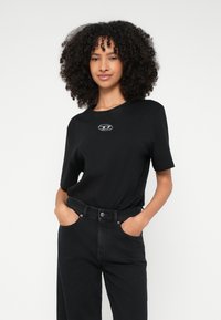 Νεαρή γυναίκα με σγουρά μαλλιά που φοράει μαύρο μπλουζάκι με ένα μικρό ασημένιο έμβλημα και μαύρο τζιν ψηλόμεσο, έχοντας τα χέρια στις τσέπες.