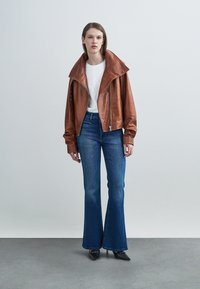 Καφέ δερμάτινο μπουφάν με φαρδιά γιακά, συνδυασμένο με λευκό t-shirt και φαρδιά τζιν παντελόνια. Μαύρα μυτερά παπούτσια ολοκληρώνουν την εμφάνιση.