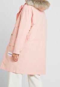 Różowy, długi płaszcz parka z kapturem wyściełanym futrem. Posiada boczne kieszenie i regulowany sznurek. Gładka tekstura i lekki materiał.