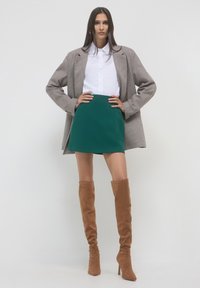 Blazer a quadre marroni sopra una camicia bianca, abbinato a una mini gonna verde e stivali al ginocchio in camoscio marrone. Texture morbide e vestibilità sartoriale.