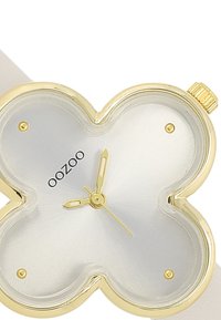 Montre dorée avec un boîtier en forme de trèfle, cadran argenté, aiguilles dorées et finition lisse. Présente un bracelet blanc et un design minimaliste.