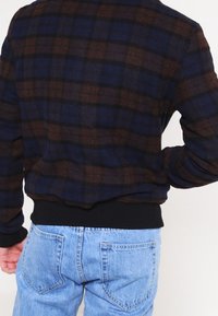 Henkilö, jolla on päällään tummansininen ja ruskea ruutujakku, jossa on mustat hihat, ja vaaleansiniset farkut, kuvattu takaa.