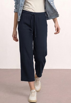 Persona che indossa pantaloni cropped a gamba larga blu navy con coulisse in vita, sneakers bianche, top bianco e giacca di jeans.