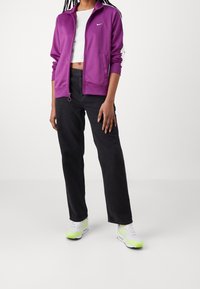 Person som bär en lila jacka med dragkedja, vit kort topp, svarta högmidjade jeans och vita sneakers med neongröna detaljer.