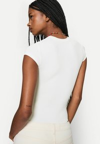 Tricou alb, ajustat, cu mâneci scurte, textură netedă și decolteu rotund. Prezintă un tatuaj vizibil pe brațul stâng.