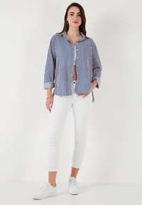 Camicia a righe blu e bianca, abbinata a jeans bianchi aderenti e sneakers bianche. Presenta una tasca e maniche arrotolate.