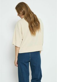 Bežový cropped top so širokými rukávmi, hladkou textúrou a voľným strihom, skombinovaný s voľnými tmavomodrými denimovými nohavicami. Zobrazený zadný pohľad.