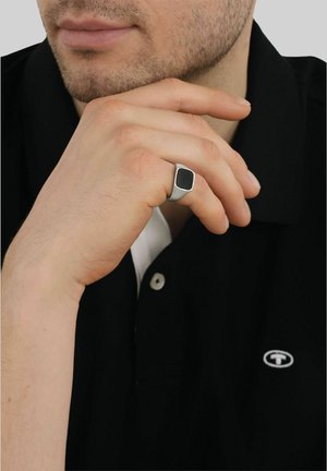 Mann mit leichtem Gesichtsstoppelbart, der ein schwarzes Poloshirt trägt und das Kinn auf eine Hand mit einem silbernen Ring mit schwarzem quadratischem Stein stützt.