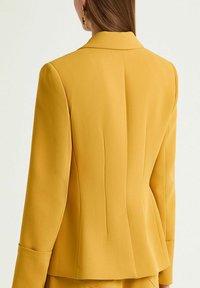 Veste jaune cintrée avec une silhouette structurée, manches longues et fente au dos. Texture de tissu lisse.
