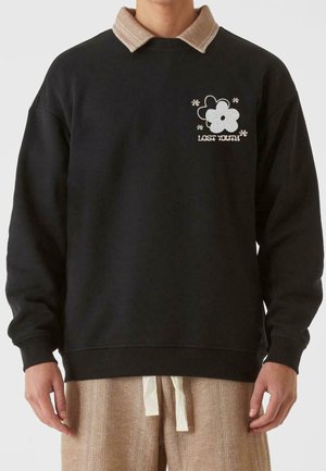 Personne portant un sweat-shirt noir avec un col beige, un motif floral et le texte "LOST YOUTH" sur la poitrine, associé à un pantalon beige texturé.