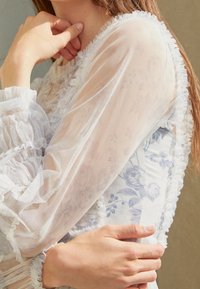 Superposition blanche transparente et froncée avec un motif floral ; design ajusté avec des accents bleu clair ; tissu texturé avec des bords doux et volantés.