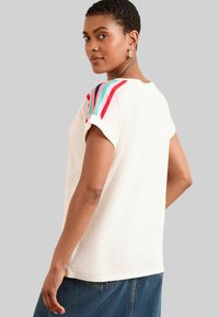 Šviesiai kreminė marškinė su trumpomis rankovėmis ir daugiaspalvėmis juostelėmis ant pečių. Minkšta tekstūra, laisvas kirpimas ir apvalus iškirpimas.