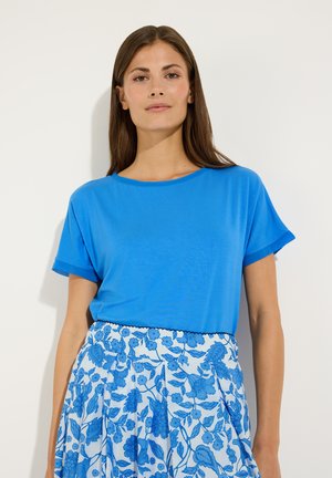 Frau mit langen braunen Haaren, die ein hellblaues kurzärmeliges Oberteil und einen weißen Rock mit blauen Blumenmustern trägt, steht vor weißem Hintergrund.