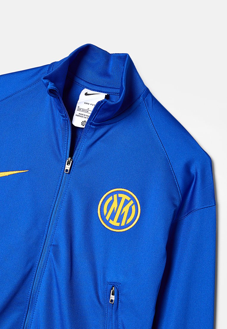 Blaues Zip-Up-Jackett mit hohem Kragen, mit gelbem Nike-Swoosh und besticktem kreisförmigem Logo mit Initialen. Glattes, leichtes Material.