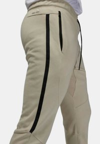 Beiga joggers med svarta sidostraper, tillverkade av mjukt, strukturerat tyg. Har midja med dragsko och dragkedjefickor på sidorna.