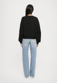 Mulher com cabelo escuro e longo, vestindo um moletom preto e jeans azuis claros com um padrão floral rosa, de pé de costas em fundo branco.