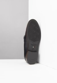 Scarpa di pelle nera con suola in gomma testurizzata che mostra motivi di grip circolari e un accento marrone sul tallone. Presenta il marchio e le indicazioni della taglia.