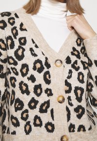 Beige vest met zwarte en beige luipaardprint, voorzien van een diepe V-hals, drie knopen en geribbelde manchetten. Zacht gebreid materiaal.