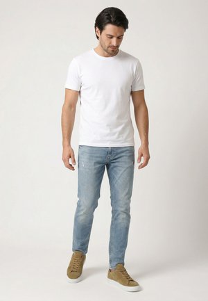 Mężczyzna ubrany w gładką białą koszulkę, jasnoniebieskie dżinsy i brązowe trampki stojący na jednolitym jasnym tle.