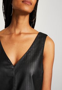 Femme aux cheveux tressés portant de grandes boucles d'oreilles en forme d'anneaux et un top noir en cuir sans manches à col en V sur un fond clair.