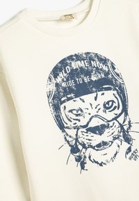 Krämfärgad sweatshirt med en marinblå grafik av en tiger i pilots hjälm. Texten lyder "VILD TID NU" och "ÅK FÖR ATT FÖDAS."