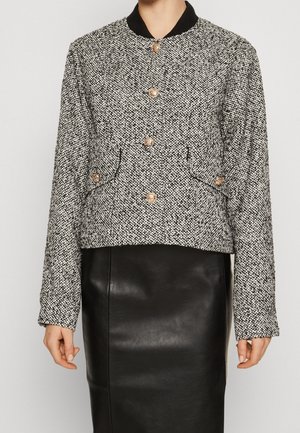 Vrouw draagt een zwart-wit tweed jasje met gouden knopen over een zwarte leren rok, staand tegen een effen achtergrond.
