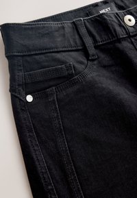 Primo piano di jeans denim neri che mostra la tasca frontale, l'asola per la cintura, il bottone argento e l'etichetta parziale del marchio con la scritta "NEXT" in vita.