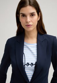 Blazer navy con spalle strutturate sopra una maglietta a righe bianco e azzurro con testo ricamato nero. Texture del tessuto liscia.