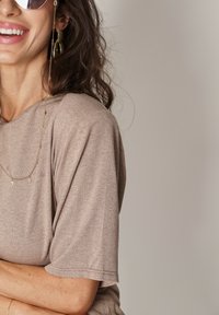 Beżowa bluzka z krótkim rękawem o luźnym kroju, z okrągłym dekoltem i gładką teksturą. Widoczny złoty naszyjnik z łańcuchem i kolczyki w kształcie kół.