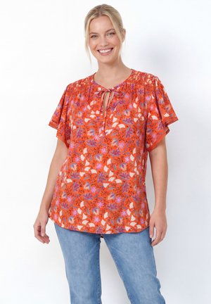 Femme souriante portant un chemisier ample orange à motifs floraux avec des manches papillon et un jean bleu sur fond blanc uni.