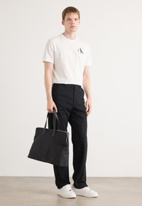 Black tote bag with dual handles, featuring a sleek design and a zippered top. The model wears a white t-shirt and black pants with white sneakers.