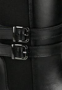 Primo piano di uno stivale in pelle nera con due fibbie regolabili in argento e pannello di tessuto testurizzato.