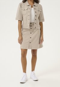 Vaaleanbeige velourtakki, jossa on lyhyet hihat, etutaskut ja nappikiinnitys. Yhdistetty vastaavaan hameeseen, jossa on nappilista. Valkoiset tennarit.