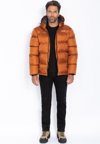Giubbotto imbottito in arancione ruggine con pattern a righe orizzontali, dettagli neri sulla cappuccio e sulla zip, abbinato a pantaloni neri e stivali beige.