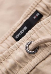 Tela beige con una etiqueta negra de ropa que dice "ambre" y un ojete metálico con un cordón beige pasado a través de él.
