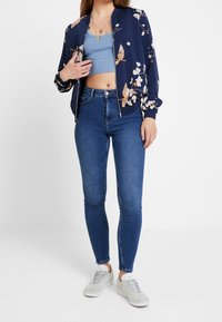 Žena v květované námořnické bundičce, modrém zkráceném tílku, úzkých džínech s vysokým pasem a světlých šedých teniskách, stojící před bílým pozadím.