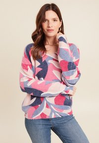 Pull en V multicolore avec un motif de feuilles roses, bleues et blanches, fabriqué en tissu doux et texturé. Porté avec un jean bleu.