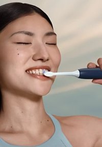 Brosse à dents électrique avec des poils blancs et des accents bleus, tenue dans la main. Une personne sourit en se brossant les dents, portant un haut bleu clair.