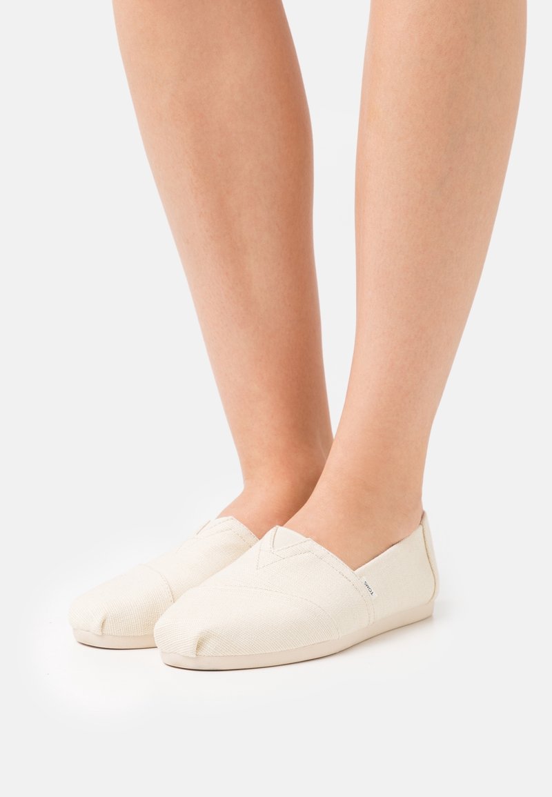 Pieds portant des chaussures en toile écrue à enfiler, avec des orteils arrondis et une couture minimale, présentés sur un fond blanc uni.