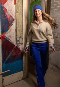 Jonge vrouw in blauwe gebreide muts, beige trui en blauwe broek loopt binnen bij een kleurrijk beschilderde houten deur en een bakstenen muur.