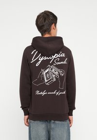 Mørkebrun hættetrøje med hvid grafik på ryggen, der viser hænder, der holder en kassetteafspiller, med tekst nedenfor.