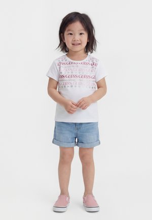 Jeune fille debout, portant un t-shirt blanc à motif graphique, un short en jean retroussé et des chaussures slip-on roses sur un fond blanc uni.