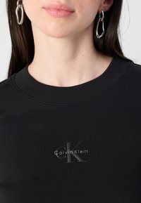 Kadar blizu osobe, prikazuje vrat i liniju čeljusti, dok nosi crni rebrasti top s Calvin Klein logotipom. Velike srebrne naušnice dopunjuju izgled.